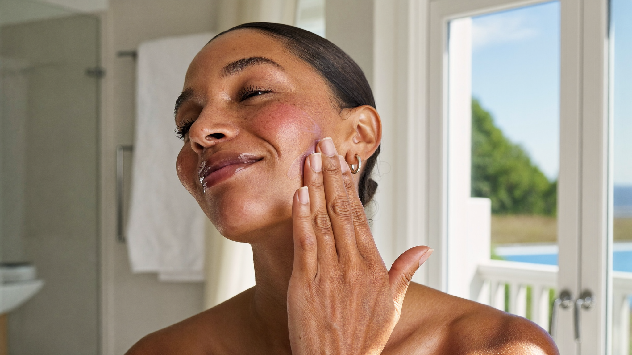 Adult woman with fresh post-treatment skin applying broad-spectrum sunscreen to her cheek in a bright bathroom as part of chemical peel aftercare